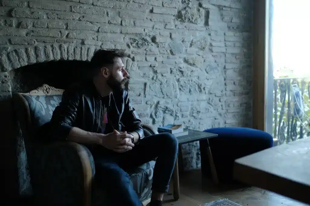 Un homme barbu d'une trentaine d'annees assis dans un fauteuil contre un mur de pierre, mains jointes, le regard tourne vers la lumiere de la fenetre