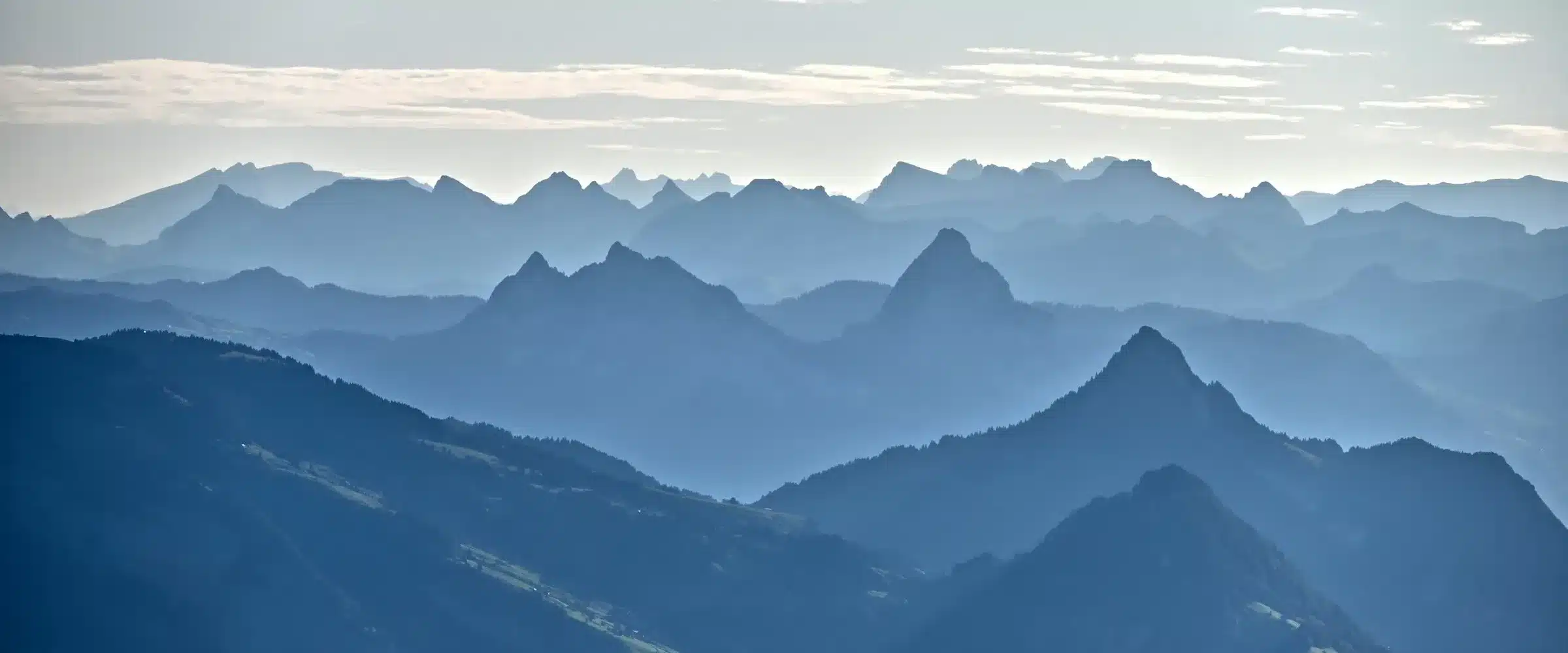 Des crêtes de Préalpes se superposent en plans successifs, du bleu profond au bleu pâle, sous un ciel clair barré d'une ligne d'horizon nette.