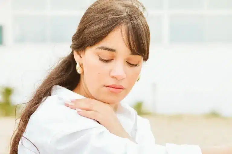 Personne portant la main à son épaule et sa nuque, près d’une fenêtre