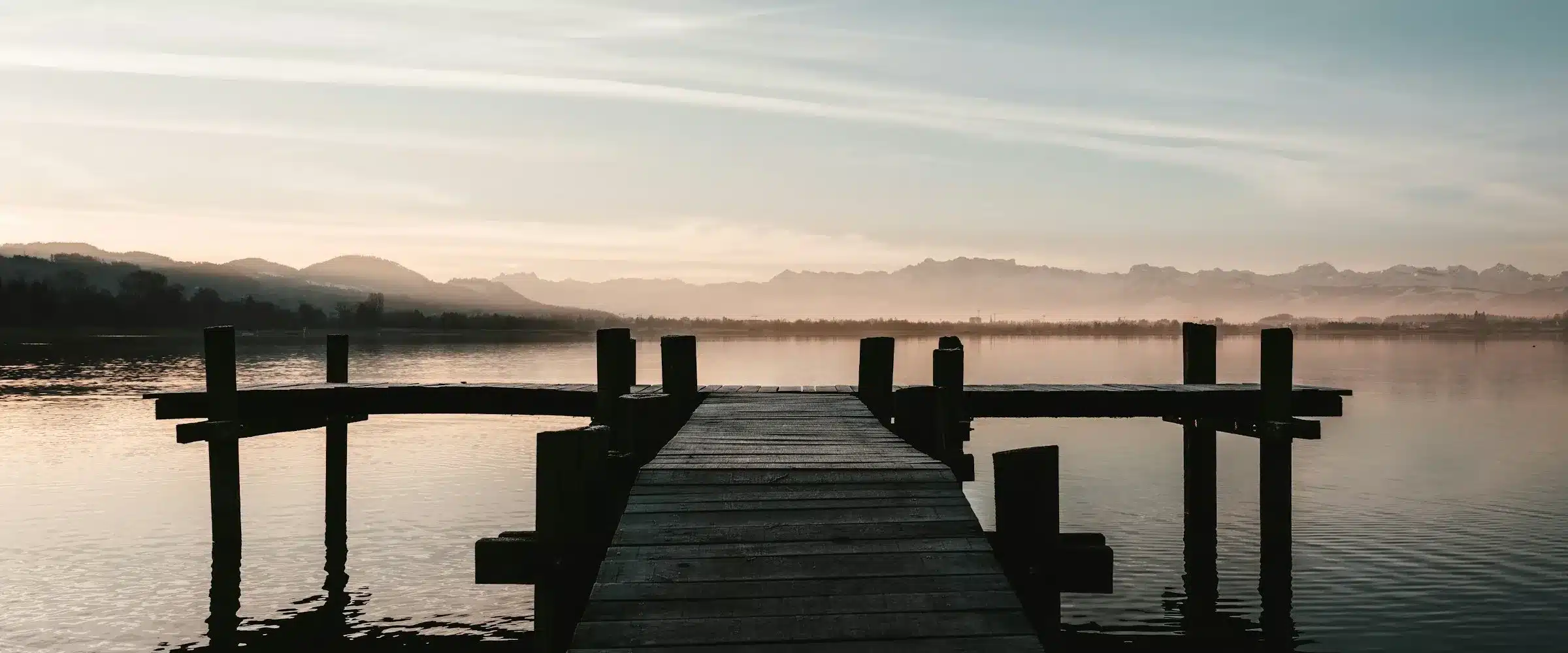 Un ponton de bois vide s'avance sur un lac immobile à l'aube ; à l'horizon, la chaîne des Alpes se découpe dans une lumière dorée.