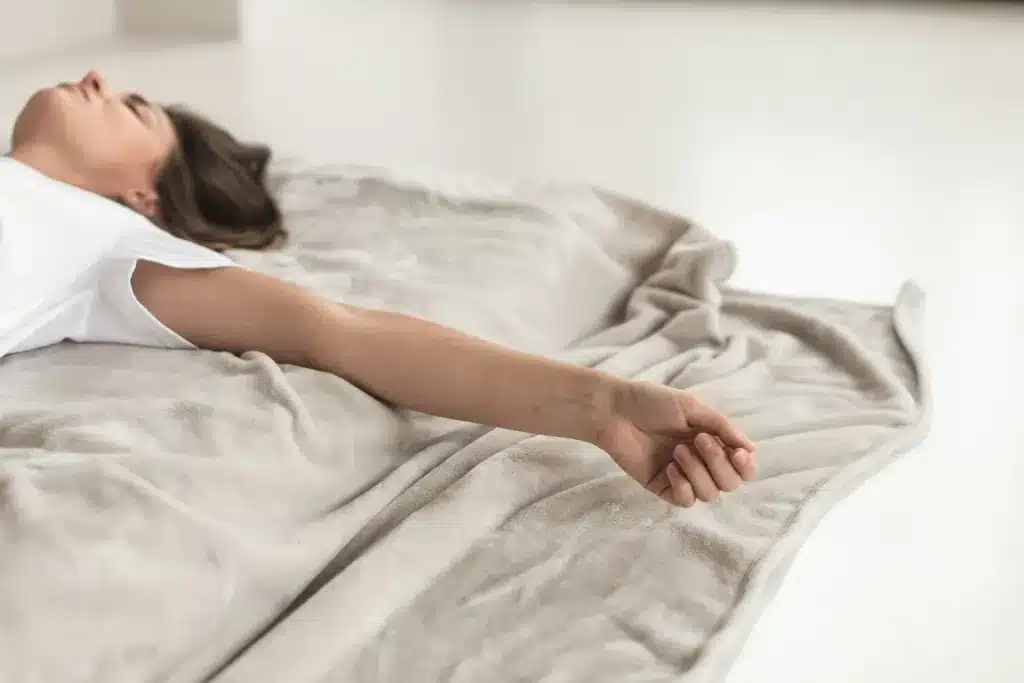 Une femme allongée sur le dos en t-shirt, recouverte d'une couverture, les yeux fermés, le bras détendu et la main ouverte, dans une lumière douce d'intérieur