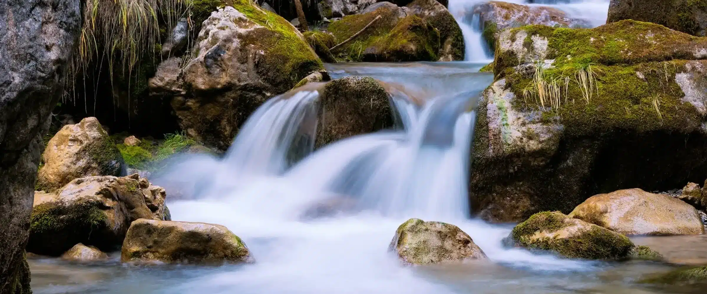 Un ruisseau de montagne descend en petites cascades entre des blocs de roche couverts de mousse ; l'eau, saisie en pose longue, se fait soyeuse et laiteuse ; touffes d'herbe sèche et lumière basse, sans ciel ni horizon.