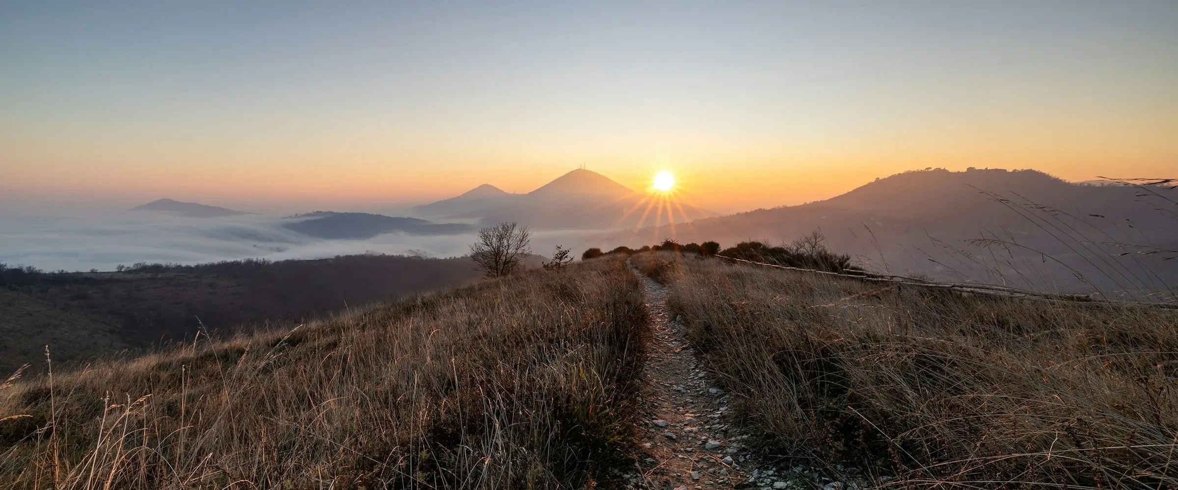 Un sentier de pierres monte le long d'une crête herbeuse vers le soleil qui se lève ; en contrebas à gauche, une nappe de brouillard remplit encore la vallée.