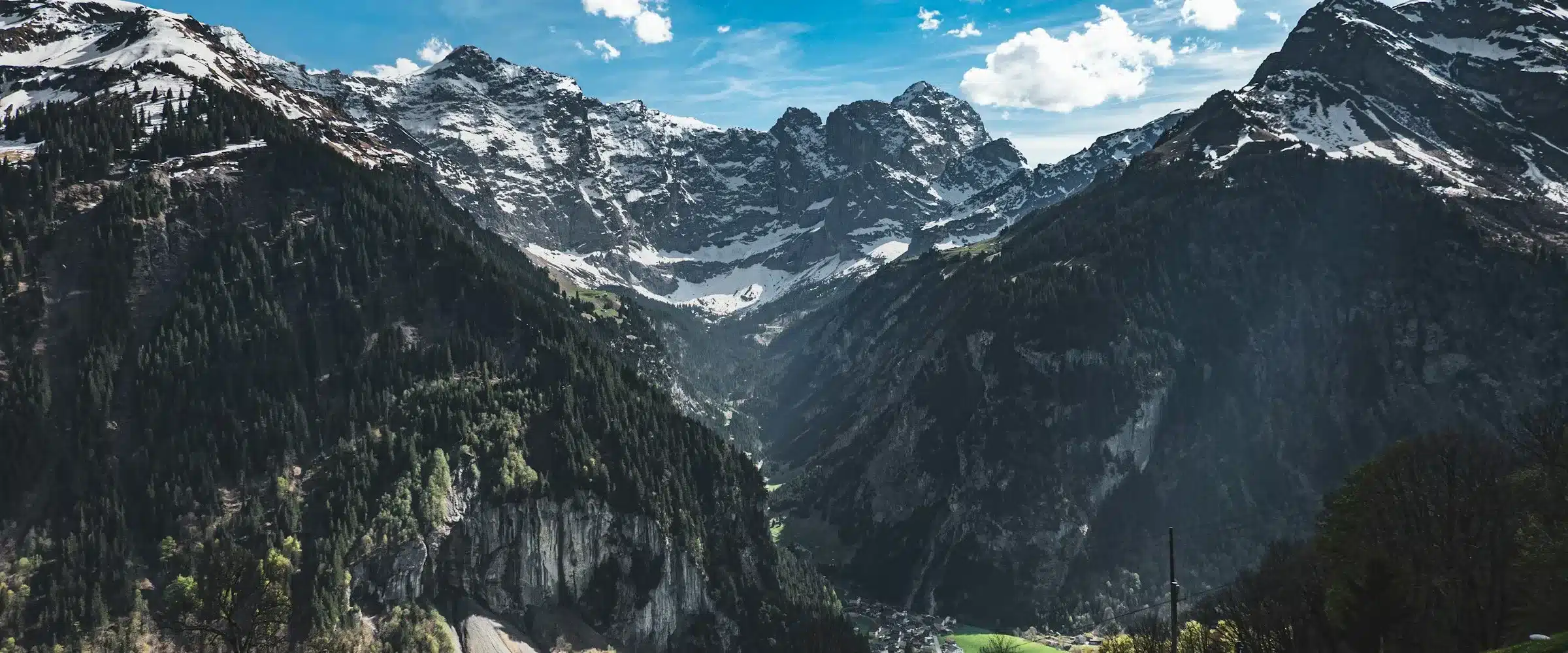 Une vallée alpine suisse en plein jour : prairie verte au premier plan, versants boisés, sommets enneigés et ciel bleu parsemé de nuages.